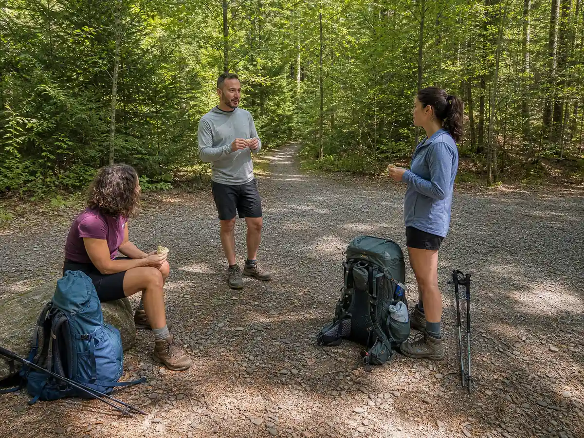 Backpacking group resting with packs and poles contained on a durable gravel turnout while the generic trail and junction remain open - considerate trail break | KnowOutdoor