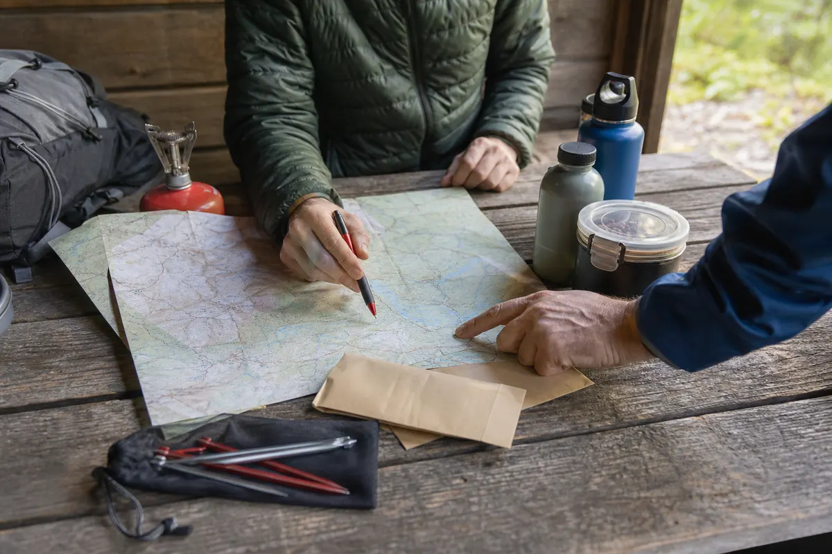 Backpackers comparing a generic official camp map, permit notes, food storage container, and water plan before choosing mileage - backpacking campsite planning | KnowOutdoor