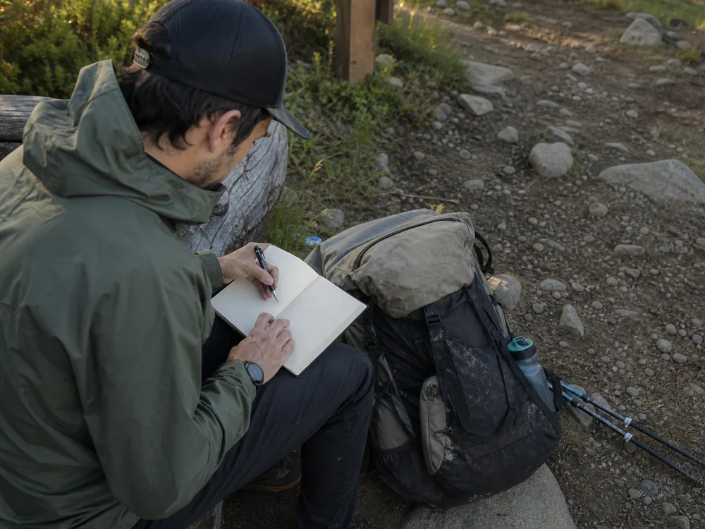 Backpacker recording moving time pack load and recovery notes beside a worn overnight pack - backpacking exercise log | KnowOutdoor