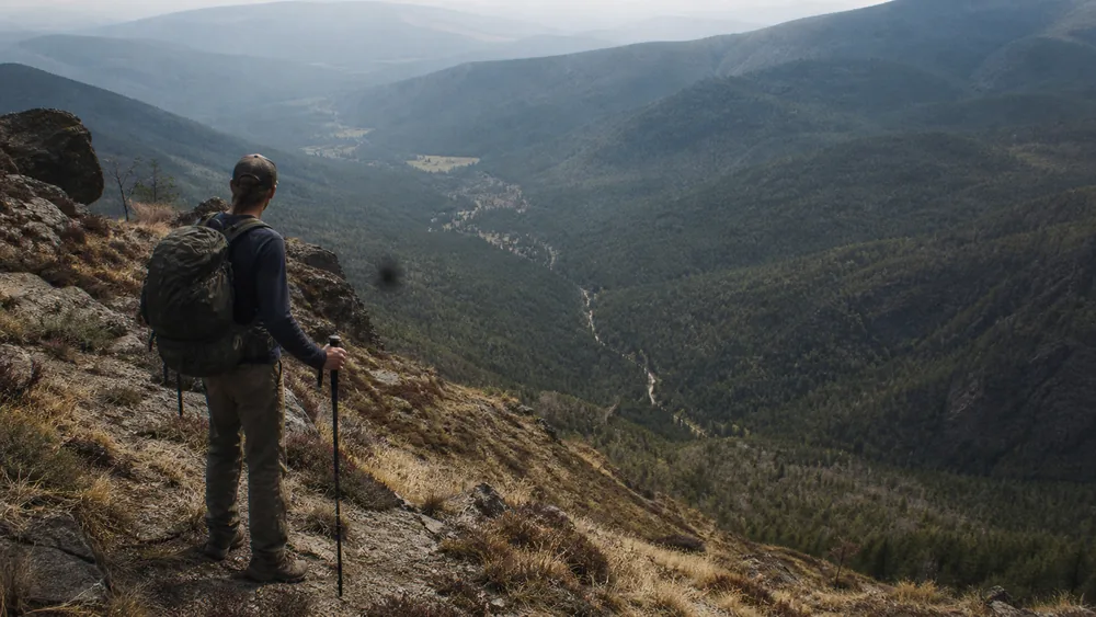 A walker on a high open ridge looking back down a long forested valley with a thin trail winding toward a distant clearing - how far the trailhead gets on an overnight trip | KnowOutdoor
