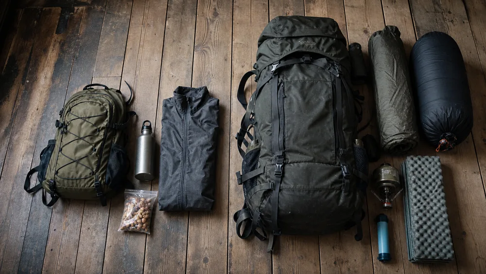 Overhead comparison of two kits on floorboards, a small daypack with bottle and jacket beside a large pack with tent, sleeping bag, mat, stove and filter - what changes between hiking and backpacking | KnowOutdoor