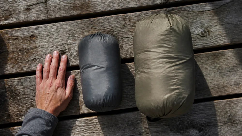 Overhead view of two stuffed sleeping bag sacks side by side on bare boards, one compressed to roughly half the size of the other, with a hand beside them for scale | KnowOutdoor