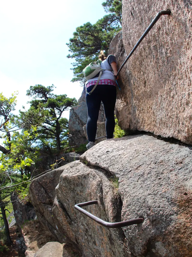 Hiker using iron rungs on an exposed granite trail in Acadia National Park - Acadia National Park | KnowOutdoor