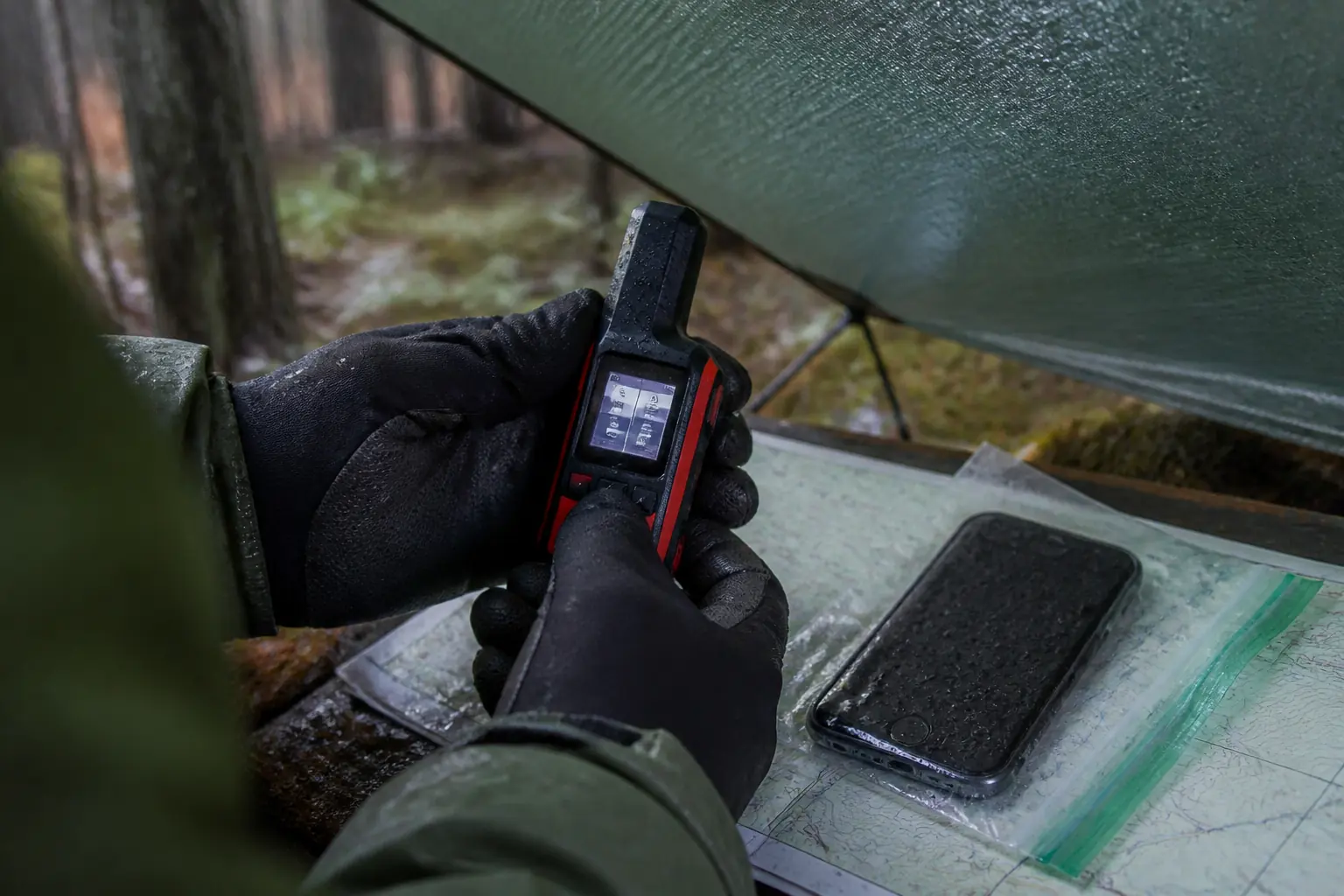 Gloved hands operate an inReach Mini 2 under a rain tarp beside a sealed wet phone and paper map | KnowOutdoor
