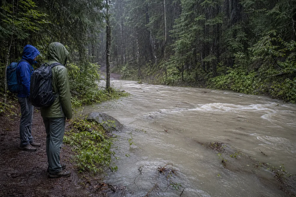 Two hikers stopped on high ground while a rain-swollen creek carries cloudy water and small debris across the route below