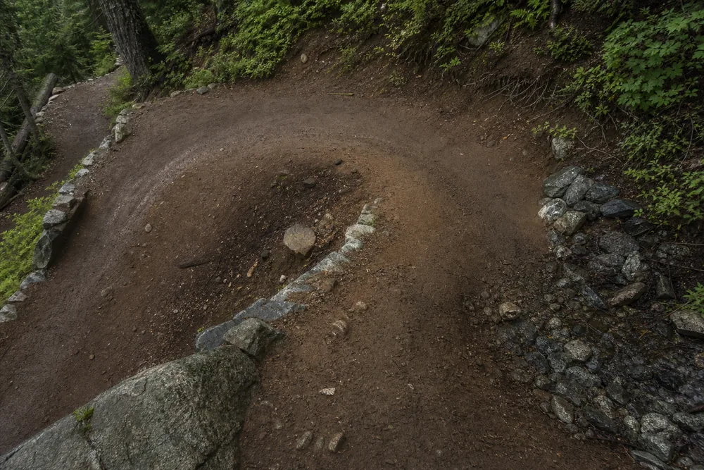Trail switchback corner seen from above with a rock-lined drainage outlet on the outer edge and a darker dished low point inside the turn - descending switchbacks | KnowOutdoor