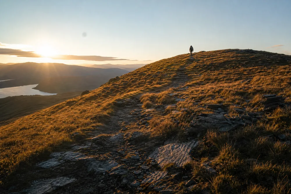 Low golden light raking across a broad ridge trail with long shadows and a single walker near the crest - sunrise and sunset hiking | KnowOutdoor