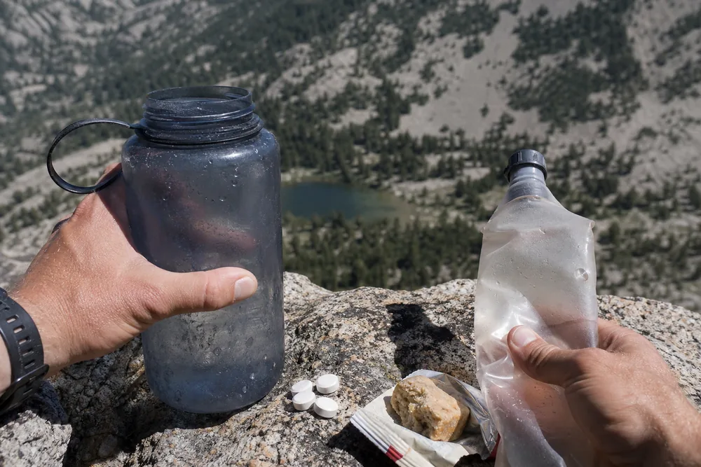 Hands holding a scratched wide-mouth bottle and a soft flask on a granite ledge above an alpine basin - hiking at altitude on a day hike | KnowOutdoor