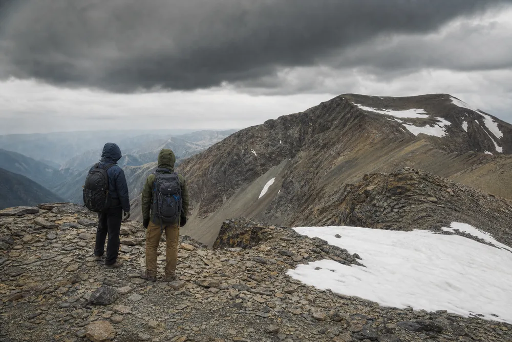 Two hikers on an exposed high saddle looking back downhill as dark storm clouds build over the ridge above - hiking at altitude on a day hike | KnowOutdoor