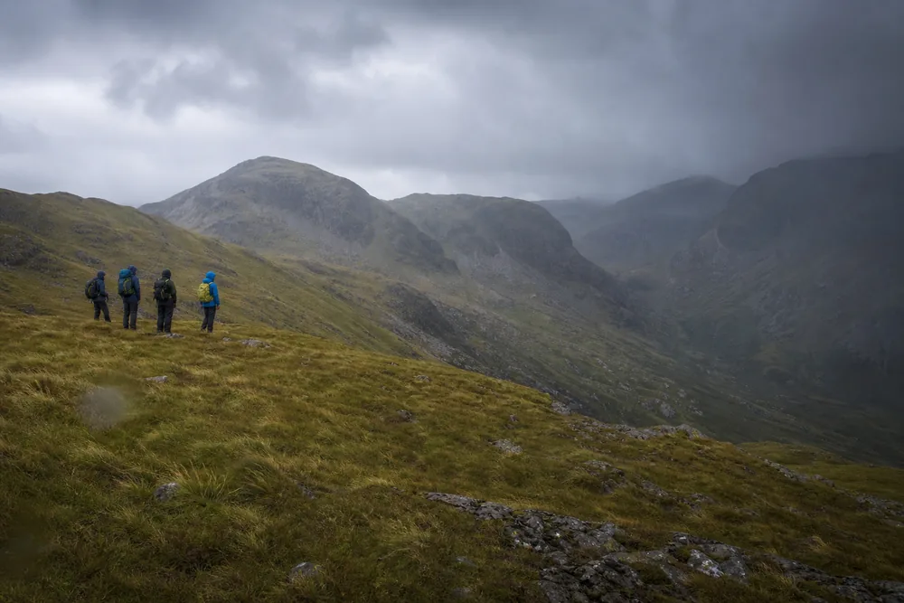Four hikers standing on an exposed grassy saddle looking back down their route as grey weather builds behind them - group hiking turnaround decision | KnowOutdoor