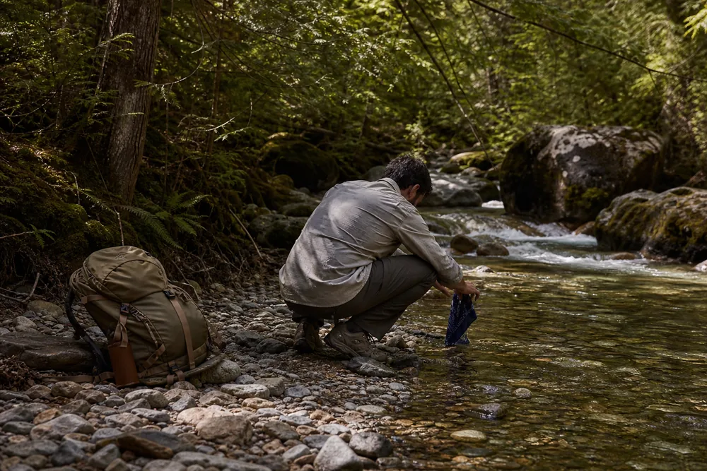 Hiker crouched at a shallow stream rinsing a bandana with a backpack resting on the bank - hiking chafing prevention | KnowOutdoor