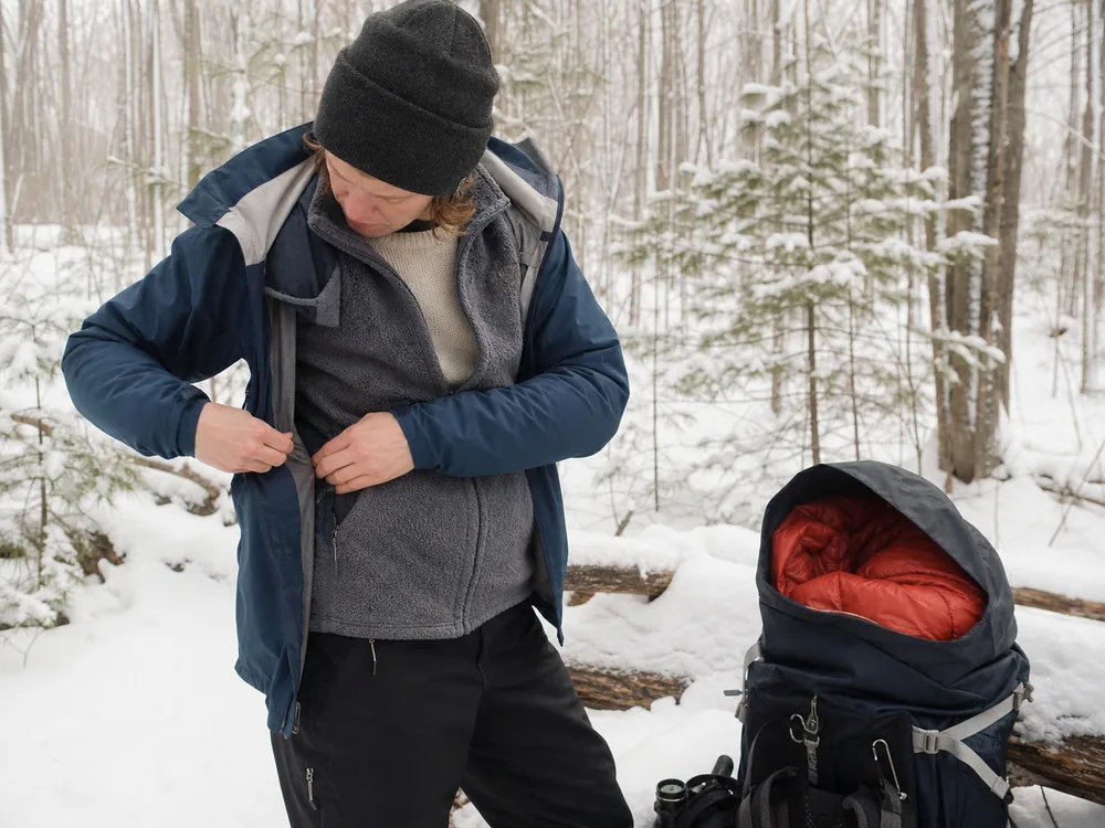 Winter hiker adjusting a shell vent with base layer midlayer and dry insulated jacket visible beside an open pack - winter hiking layer system | KnowOutdoor