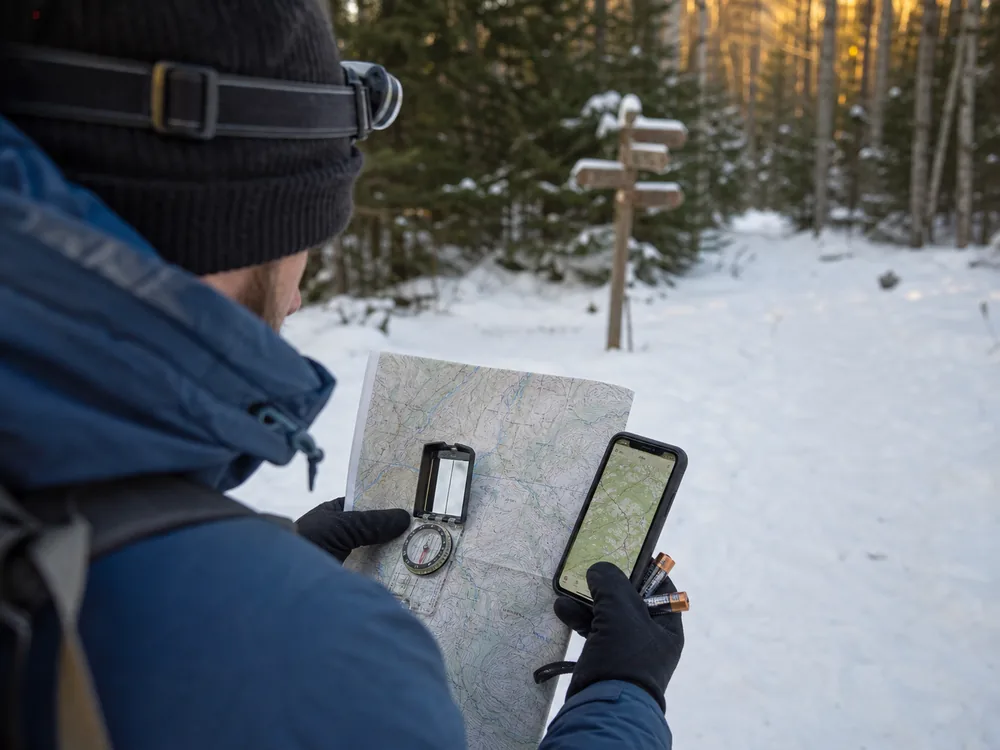 Gloved hands comparing a paper topographic map compass offline phone map and headlamp on a snowy log - winter hiking navigation kit | KnowOutdoor