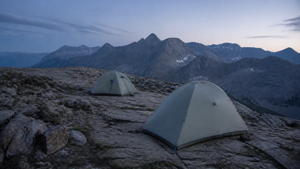 Small tents pitched on a bench above treeline with high ridges behind in flat evening light - high sleeping elevation and altitude sickness risk | KnowOutdoor