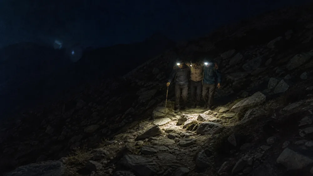 Headlamp beams on a rocky trail as two walkers help a third descend at night below a high camp - night descent decision in altitude illness | KnowOutdoor