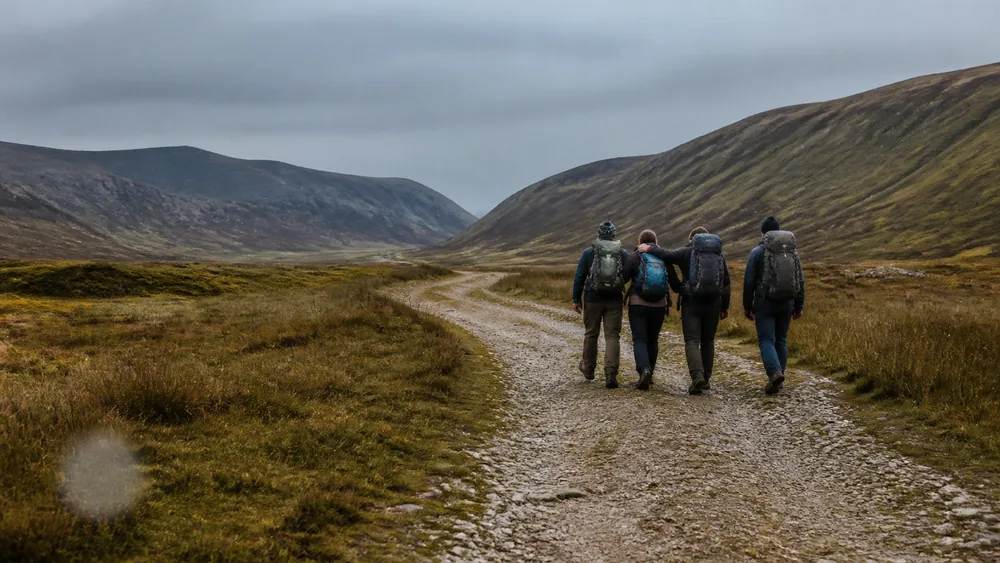 Three walkers moving steadily down a valley trail supporting a fourth between them in flat afternoon light - continuing an evacuation after an anaphylactic reaction | KnowOutdoor