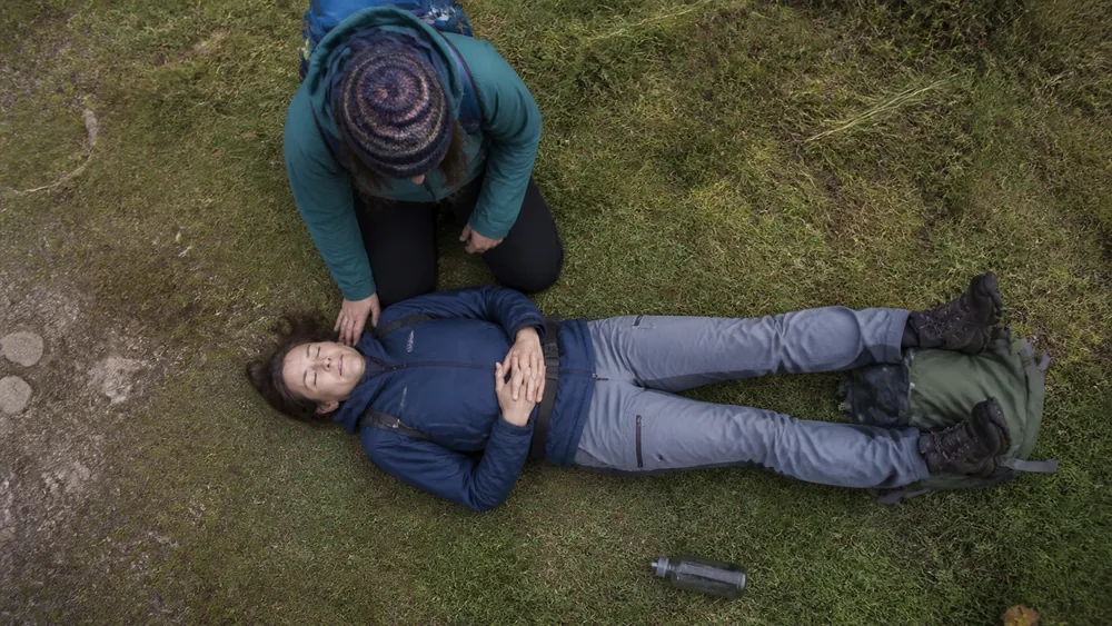 A walker lying on their back on a trail with their legs raised on a pack while a companion kneels beside them - positioning during an anaphylactic reaction | KnowOutdoor