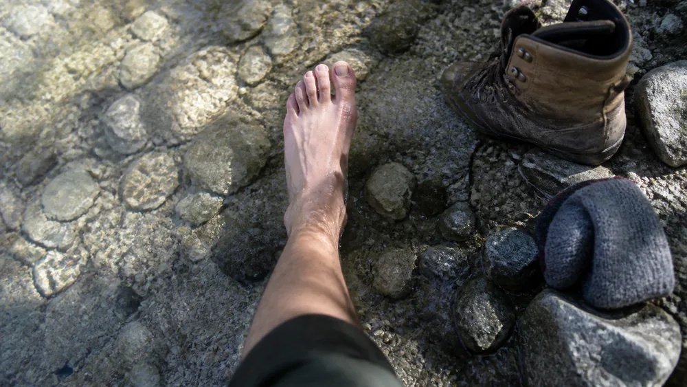 Overhead close view of a bare foot and ankle held in a shallow clear stream running over pale gravel with an unlaced boot and wool sock on the bank - cold water as field treatment | KnowOutdoor