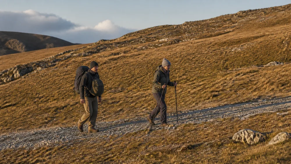 Two hikers on a broad stony track in low golden light, the front one walking with two poles and no pack while the second carries a pack on their back and another on their front - walking out an injured party member | KnowOutdoor
