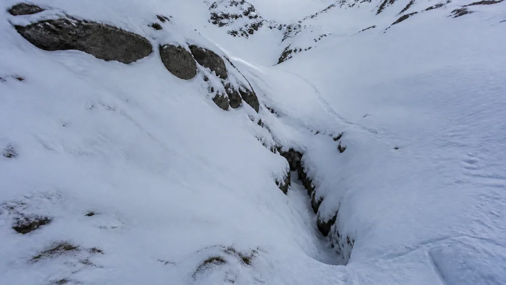 A snow-filled gully cutting steeply across a winter walking route with the packed boot track disappearing into the drainage and reappearing beyond - avalanche terrain on a hiking trail | KnowOutdoor