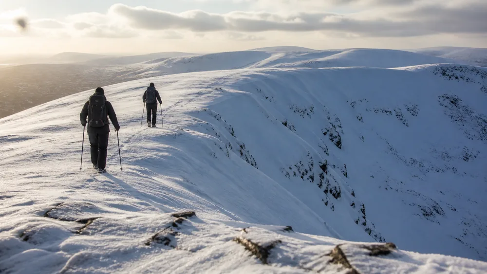 Two winter walkers following a broad rounded ridge of wind-packed snow with a steep open bowl dropping away to one side - choosing low-angle ground over the slope that could slide | KnowOutdoor