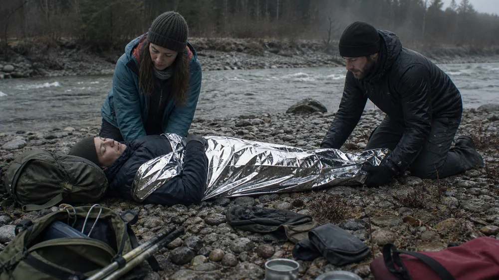A soaked walker lying flat and wrapped in a foil blanket on a river bank while two companions kneel beside keeping them horizontal | KnowOutdoor