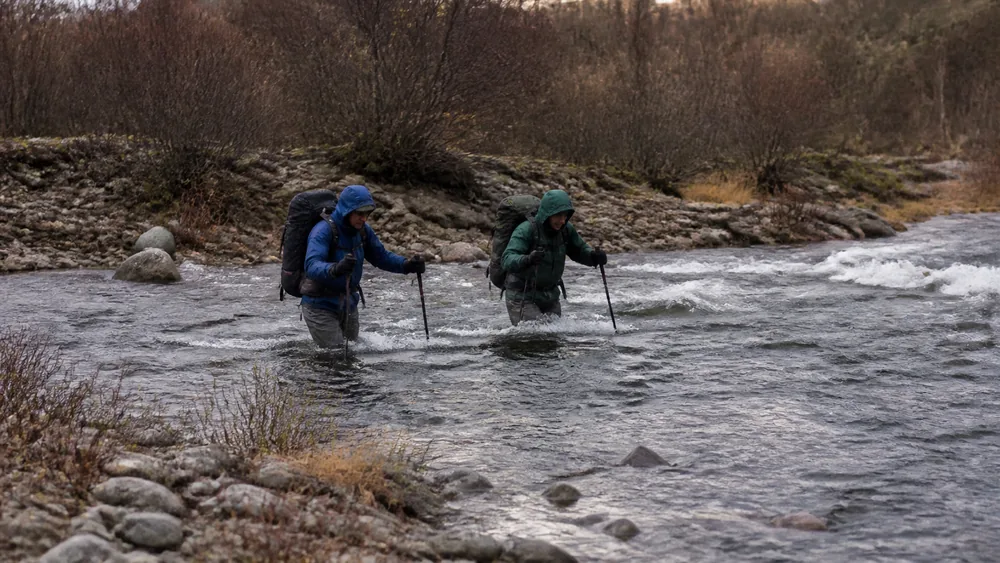 Two walkers wading a shallow fast river together with packs on and poles planted upstream, cold water breaking white around their legs | KnowOutdoor