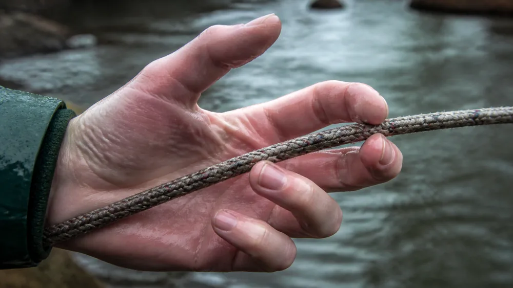 Close view of a pale reddened hand failing to close properly around a wet rope on a cold river bank, fingers stiff and curled | KnowOutdoor