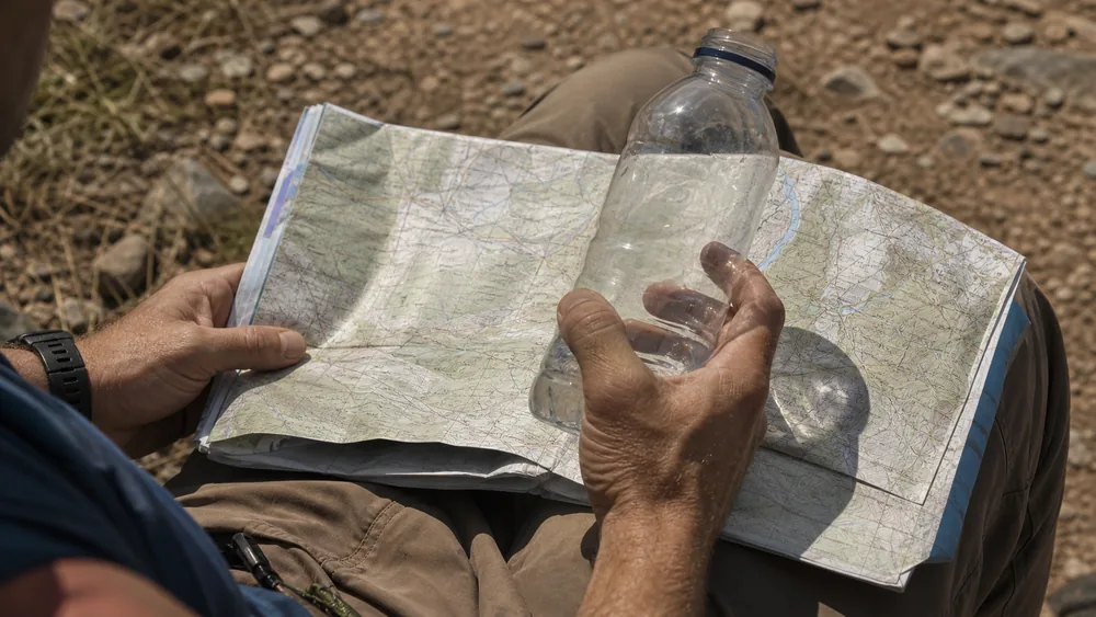 Hands holding a part-empty water bottle against a folded paper map on a walker's knee, judging what is left against the distance remaining | KnowOutdoor