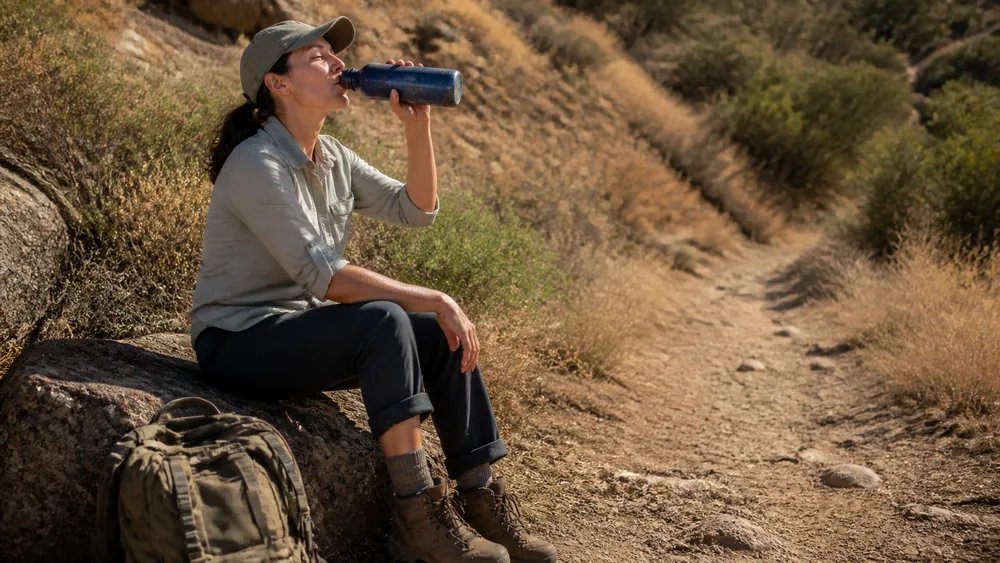 A hiker sitting on a rock at the side of a trail drinking unhurriedly from a scratched water bottle with the pack off beside them | KnowOutdoor