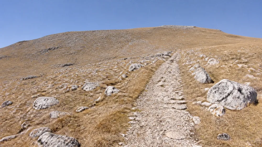 A stony trail crossing a wide open slope with no tree cover, sun high and shadows short, dry grass and pale rock stretching away | KnowOutdoor