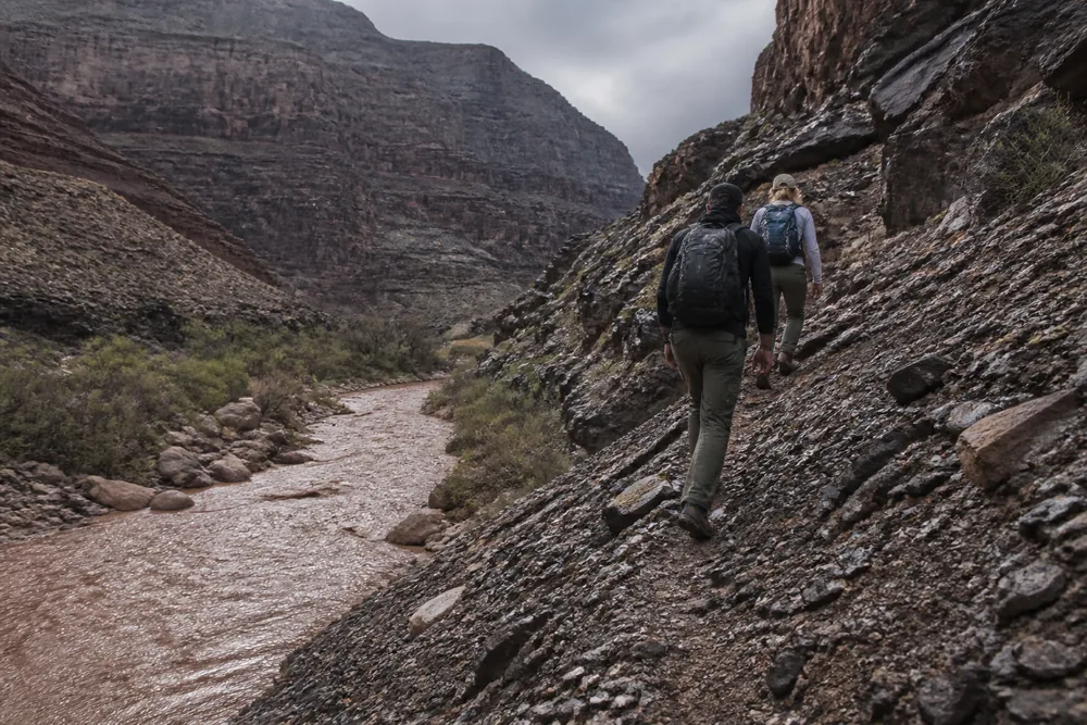 Two walkers on a steep rocky slope well above a muddy creek running through a desert canyon | KnowOutdoor
