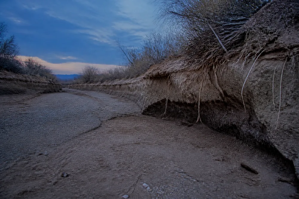 A flat sandy wash floor between two low scoured banks at dusk, bare of vegetation, with a cut bank and exposed roots on one side | KnowOutdoor