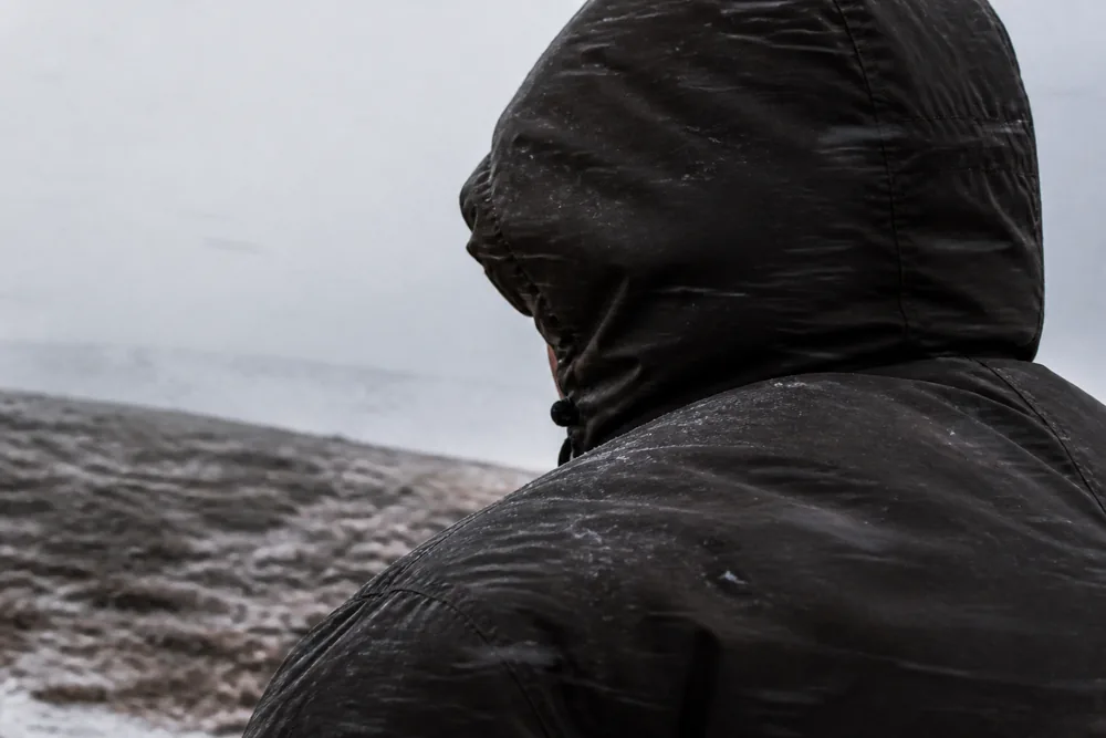 Close view from behind of a walker's hood and shoulder on a winter ridge, hood cinched across the face and frost crusted on the shell fabric | KnowOutdoor