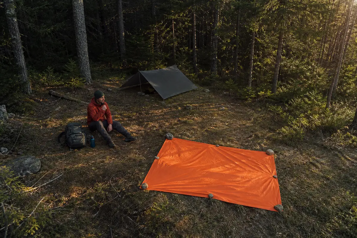 Lost hiker beside a compact shelter with a bright orange signal panel spread in a small conifer clearing - getting lost signal site | KnowOutdoor