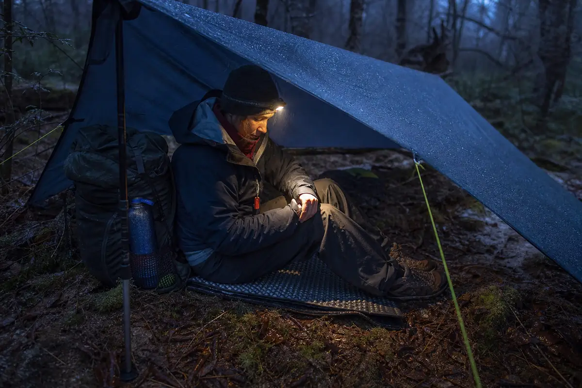 Lost hiker on an insulated pad under a low rain tarp with water and headlamp ready at dusk - getting lost waiting shelter | KnowOutdoor