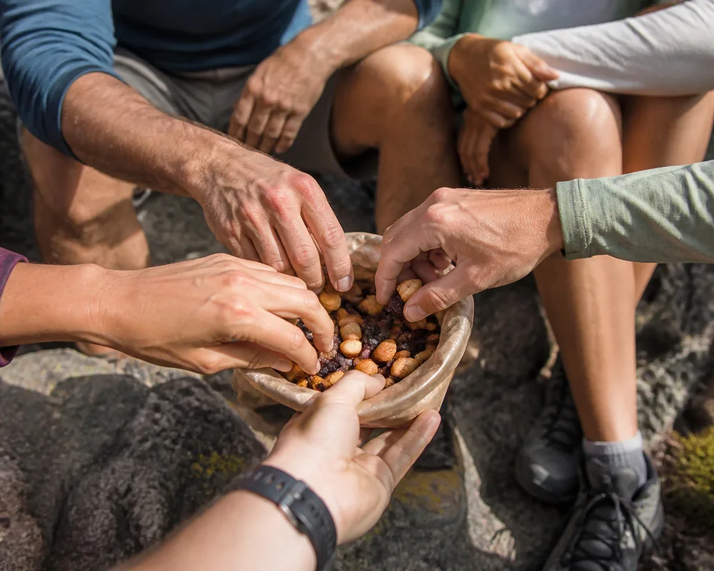 Several hands reaching into an open bag of trail mix passed around a group sitting on rocks at a rest stop | KnowOutdoor