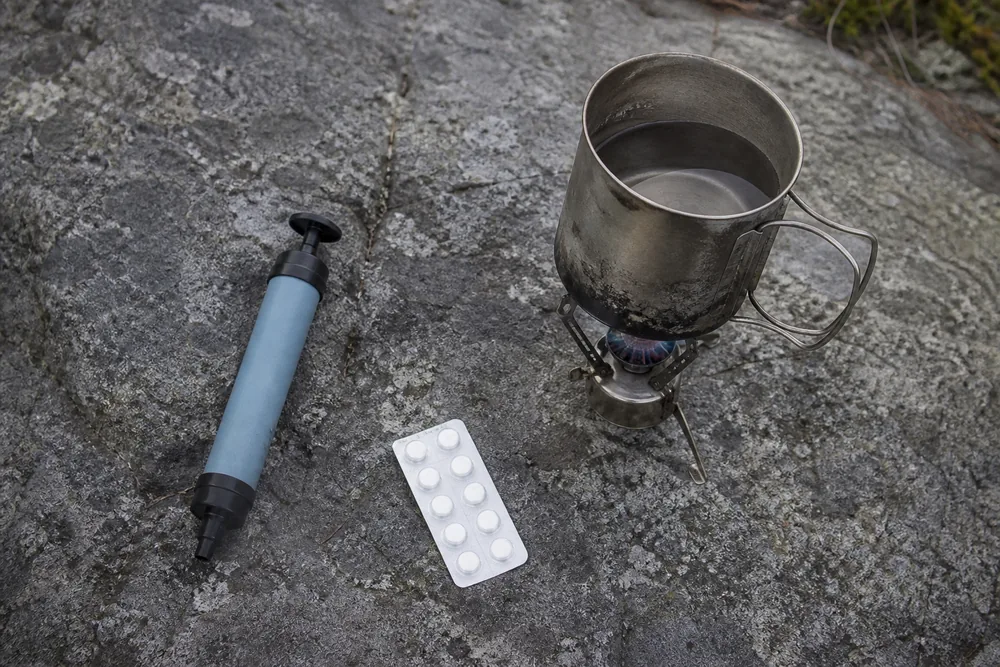 A squeeze water filter, a blister strip of purification tablets and a small pot on a stove arranged on a flat rock | KnowOutdoor