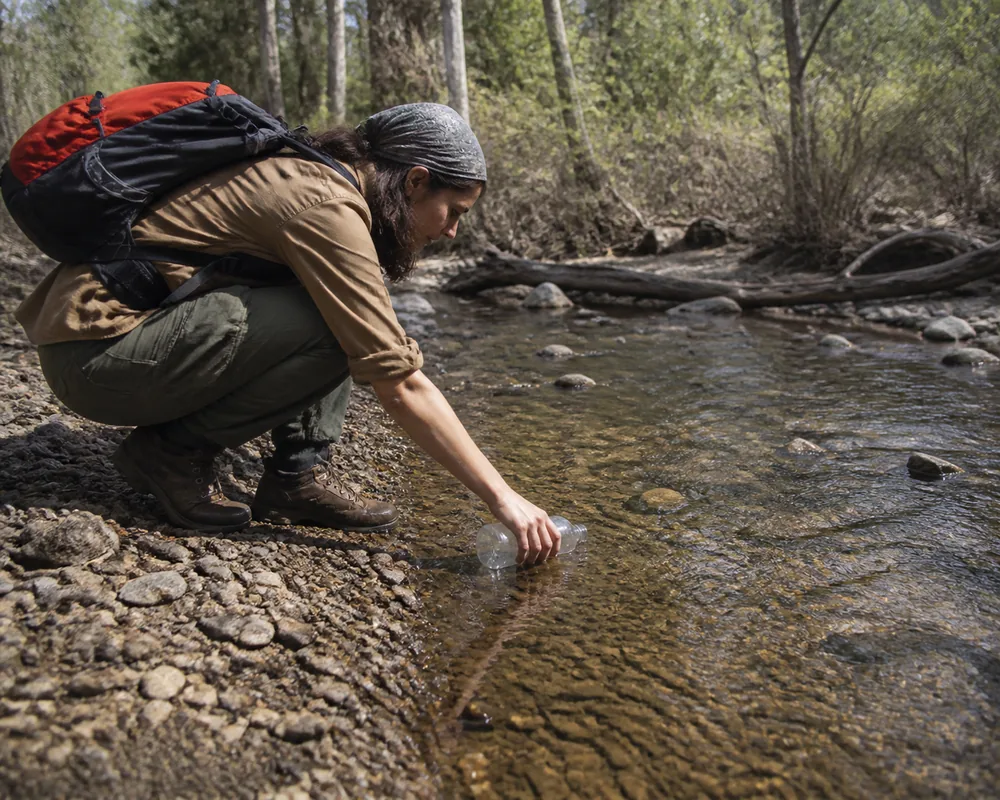 A hiker crouching to fill a bottle from a shallow slow-moving stream over gravel in open woodland | KnowOutdoor