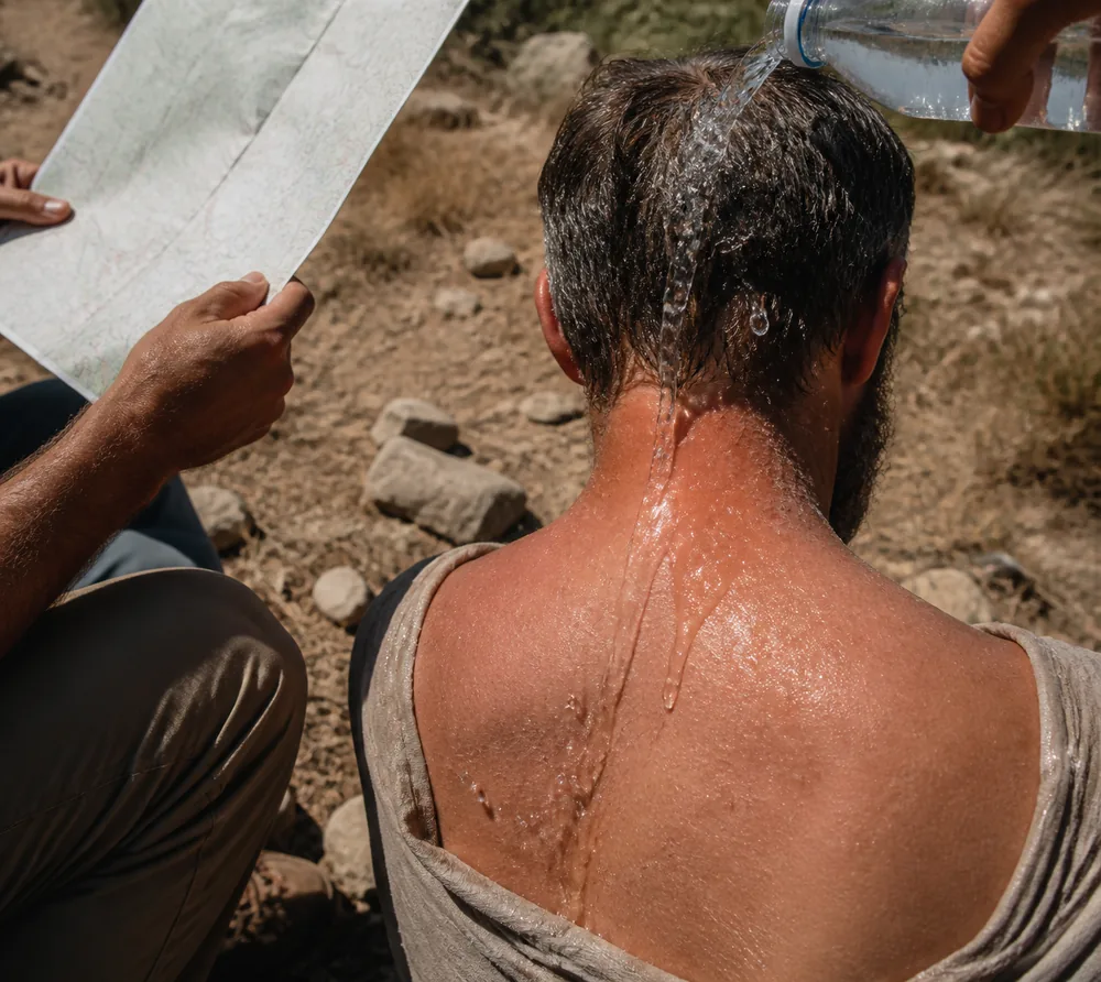 Water being poured from a bottle over the back of a seated hiker's neck while another person fans them with a folded map | KnowOutdoor