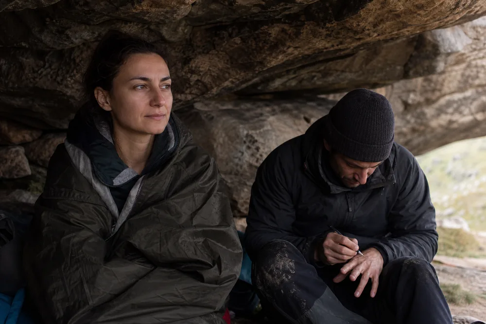 A walker sitting wrapped in a jacket in deep shade while a companion crouches beside them writing on the back of their own hand | KnowOutdoor