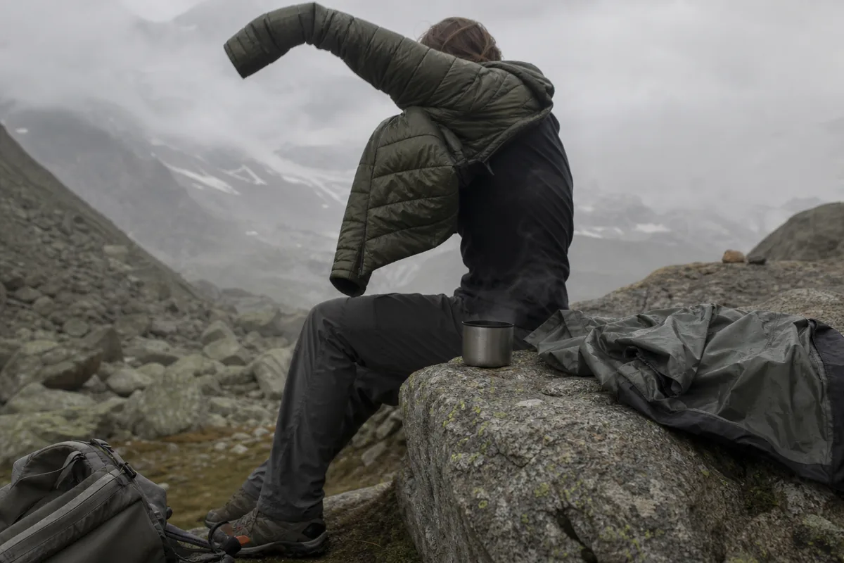 Hiker pulling on a dry insulated jacket with a hot drink at a cold sheltered stop - Hypothermia prevention | KnowOutdoor