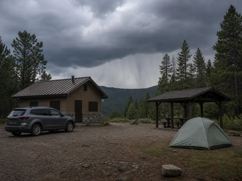 An enclosed hard-topped vehicle and substantial trailhead building contrasted with a tent and open picnic pavilion beneath dark clouds - protective and unsafe lightning shelters | KnowOutdoor