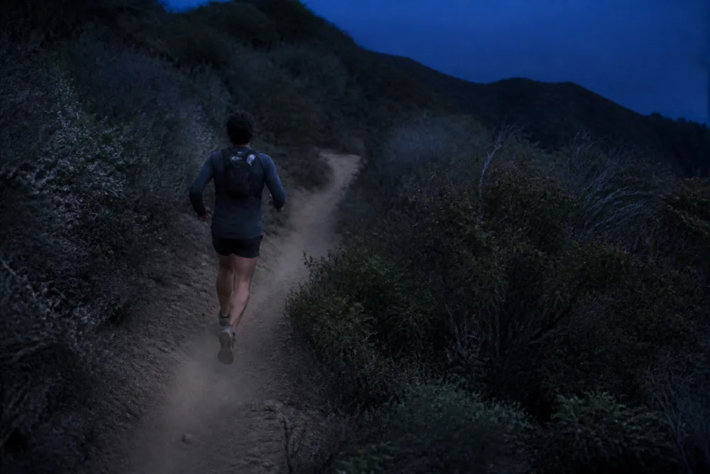 A lone trail runner on a narrow single track through dense chaparral in fading blue evening light with brush crowding the trail edge and a blind bend ahead - the speed, solitude and low light that concentrate the risk | KnowOutdoor