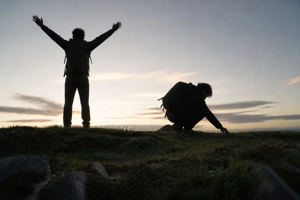 Two silhouettes of the same hiker side by side against a bright dawn sky, one standing tall with arms raised and one crouched low reaching toward the ground - how each outline reads at an animal's eye level | KnowOutdoor