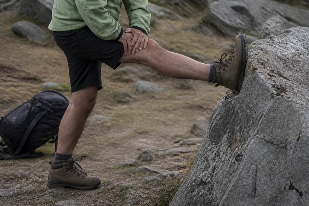 A hiker on a rocky trail with one leg straight and the ball of the foot pressed against a raised rock, ankle flexed so the toes are drawn toward the shin - the straight-knee geometry that releases a calf cramp | KnowOutdoor