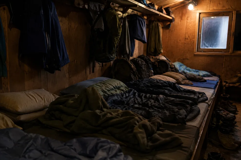 The communal sleeping room of a plain alpine hut with mattresses laid edge to edge along a continuous wooden platform, packs and damp jackets crammed against the wall and boots piled at the end | KnowOutdoor