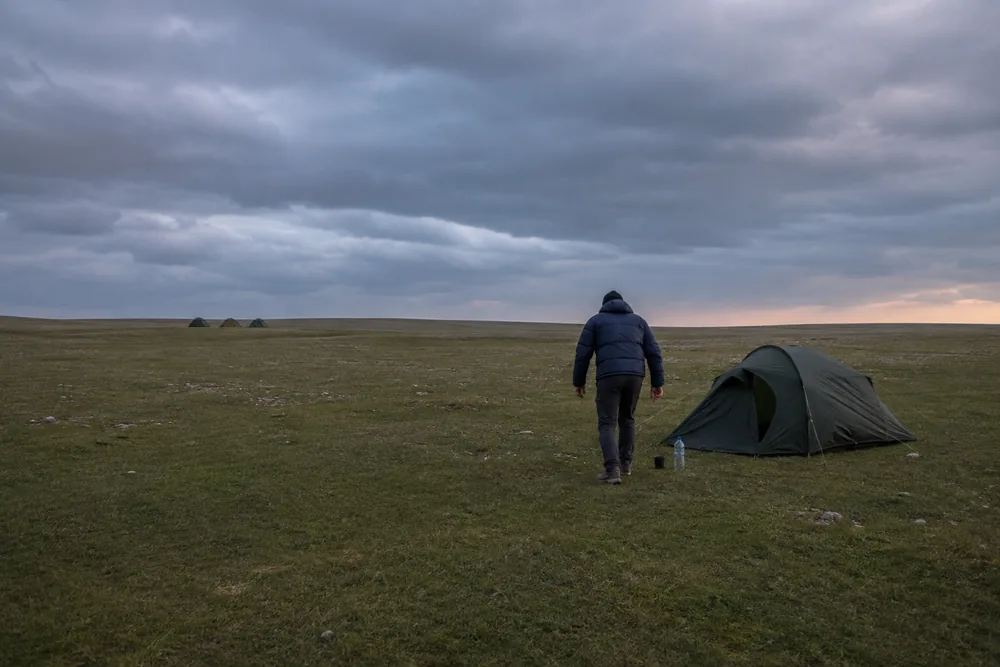 A single tent pitched far out on open grass well away from a cluster of three other tents, with one person setting a filled water bottle and a mug on the ground a couple of paces short of its door and already turning to leave | KnowOutdoor