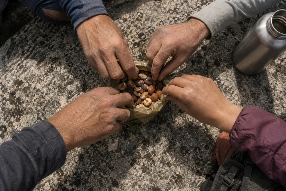 Several hikers' dusty hands reaching into one open communal snack bag on a granite slab at a rest stop, with a shared water bottle beside it - the most direct route norovirus takes between people | KnowOutdoor