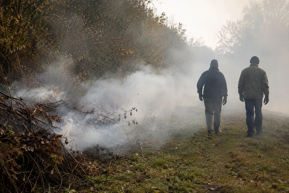 A brush clearance pile smouldering at an overgrown woodland edge with pale smoke drifting low and sideways across a grassy track, two people in work gloves walking through the drift with their backs to the camera | KnowOutdoor